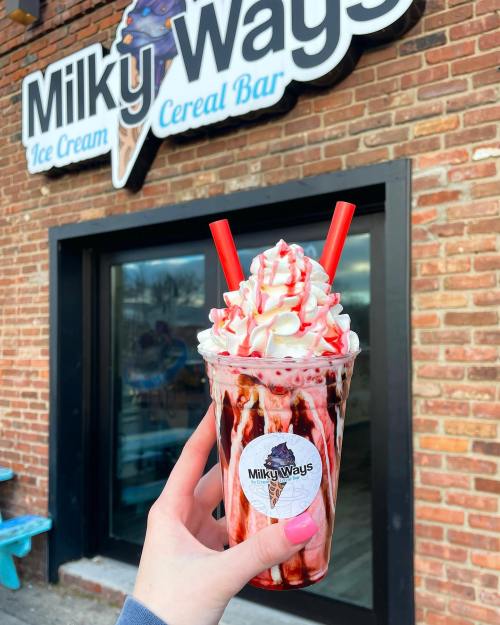 A hand holds a colorful milkshake topped with whipped cream and red drizzle, in front of a "Milky Ways" sign.