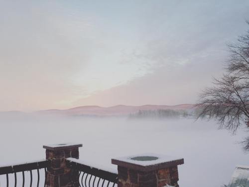 A serene winter landscape with fog over a frozen lake and distant mountains, viewed from a stone railing.