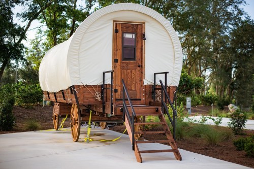 A wooden wagon with a white canvas cover, featuring a door and stairs, set in a landscaped outdoor area.