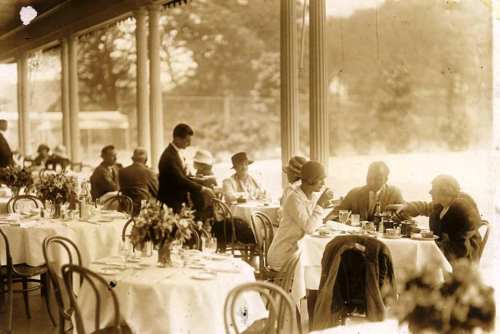 A vintage dining scene with elegantly dressed patrons enjoying meals at tables adorned with flowers.
