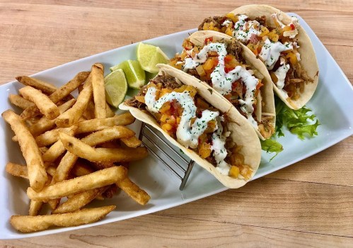 Three tacos filled with shredded meat and toppings, served with fries and lime wedges on a white plate.