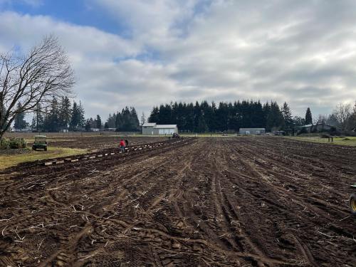 A plowed field under a cloudy sky, with workers and machinery in the distance, surrounded by trees and buildings.