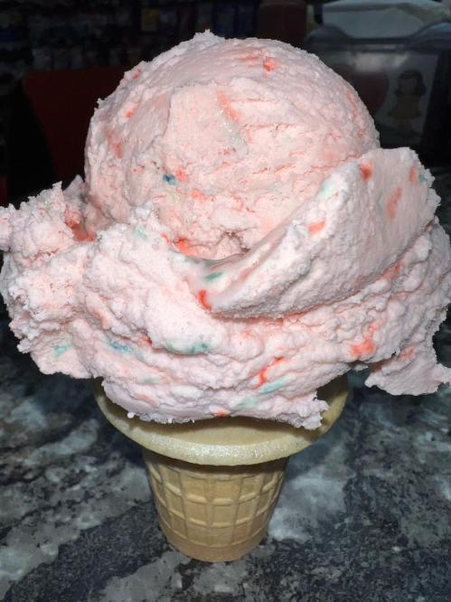 A large scoop of pink ice cream with colorful specks on a waffle cone, set against a dark countertop.