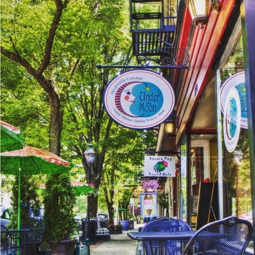 A charming street scene featuring colorful umbrellas and signs for "Under Moon" restaurant amidst lush green trees.