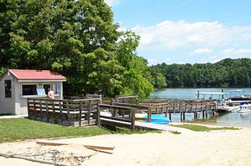 A lakeside scene featuring a small cabin, wooden docks, and boats, surrounded by trees and a sandy beach.