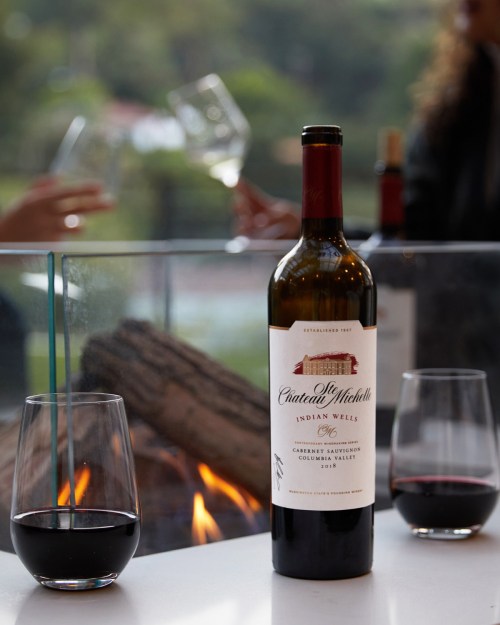 A bottle of Chateau Ste. Michelle Cabernet Sauvignon with two glasses of red wine, set near a cozy fireplace.