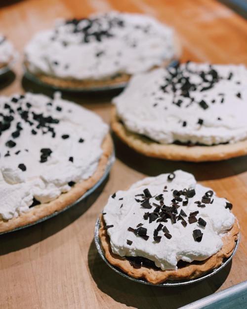 Several chocolate cream pies topped with whipped cream and chocolate shavings on a wooden surface.