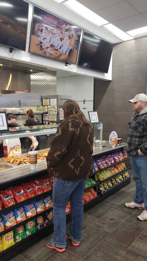A customer stands at a counter looking at food options, while another person waits nearby. Colorful snack bags line the bottom.