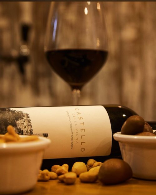A glass of red wine beside a bottle labeled "Castello" and bowls of nuts and olives on a wooden surface.