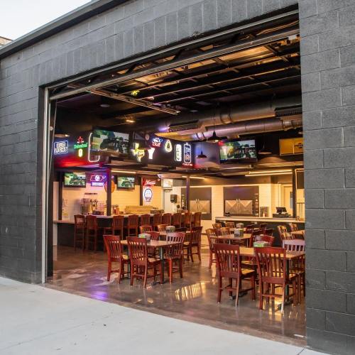 View of a restaurant interior through an open garage door, featuring wooden tables, chairs, and multiple TVs.