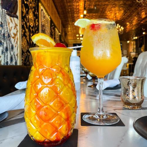 Two colorful cocktails on a marble table: a pineapple-shaped glass and a tall glass with orange drink and garnishes.