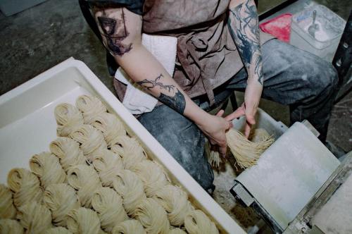 A person with tattoos prepares fresh noodles, surrounded by neatly arranged bundles of pasta in a kitchen setting.