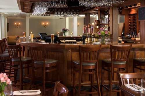 A cozy bar area with wooden stools, a polished counter, and glasses hanging overhead, surrounded by elegant decor.