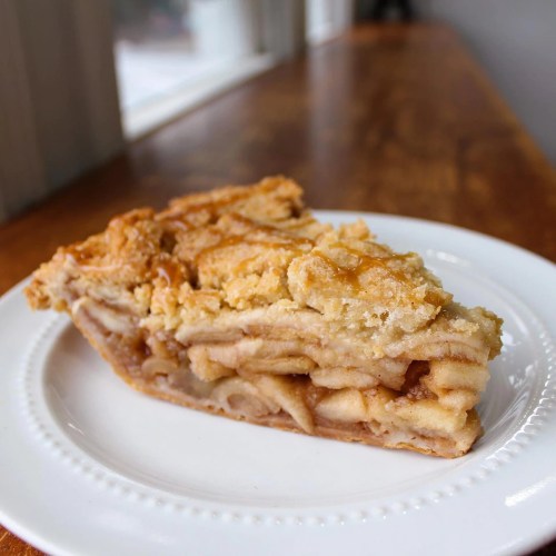 A slice of apple pie on a white plate, featuring a golden, crumbly topping and visible apple filling.