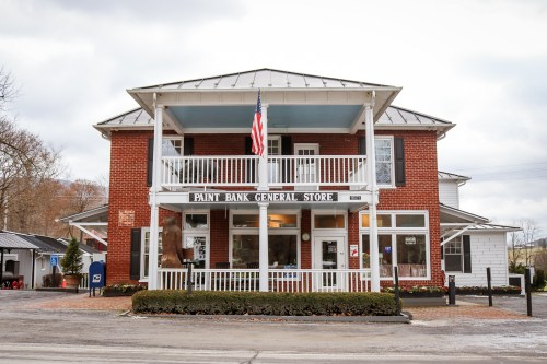 A brick general store with a porch, American flag, and large windows, surrounded by greenery and a gravel path.