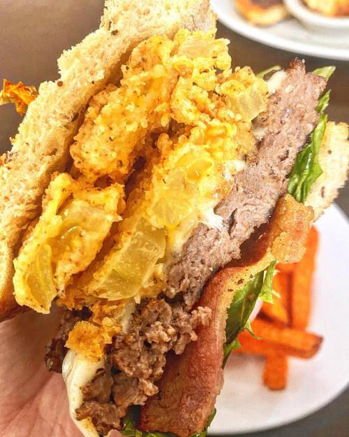 A close-up of a hearty burger with layers of beef, lettuce, crispy onion rings, and sauce, served with sweet potato fries.