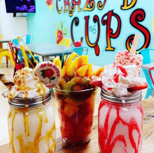 Three colorful desserts in jars: a milkshake with whipped cream, a fruit cup, and a sundae with toppings, set in a vibrant café.