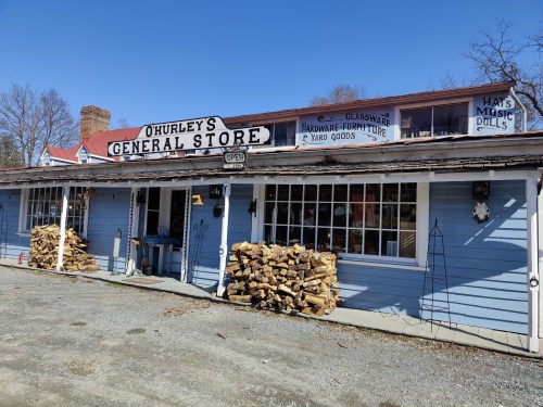 A blue general store with a sign, stacked firewood, and various goods displayed outside on a sunny day.