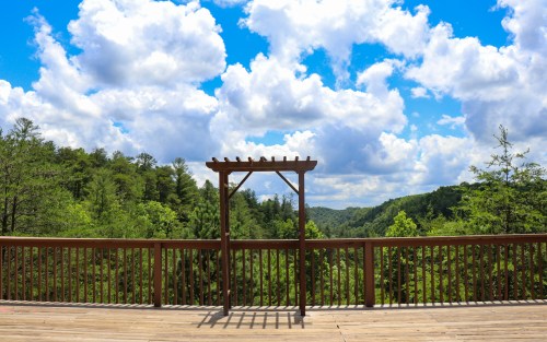 A wooden deck overlooks a lush green landscape under a bright blue sky with fluffy white clouds.