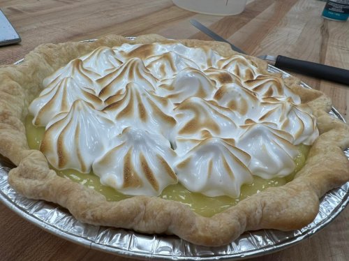 A freshly baked lemon meringue pie with golden-brown peaks on top, resting in a silver pie dish on a wooden surface.