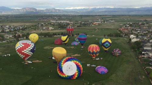 Aerial view of colorful hot air balloons on a grassy field with mountains in the background and nearby houses.
