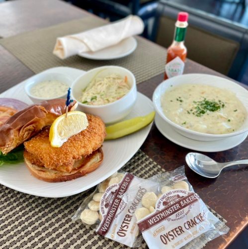 A plate featuring a fried fish sandwich, coleslaw, clam chowder, and oyster crackers, with lemon and condiments on the side.