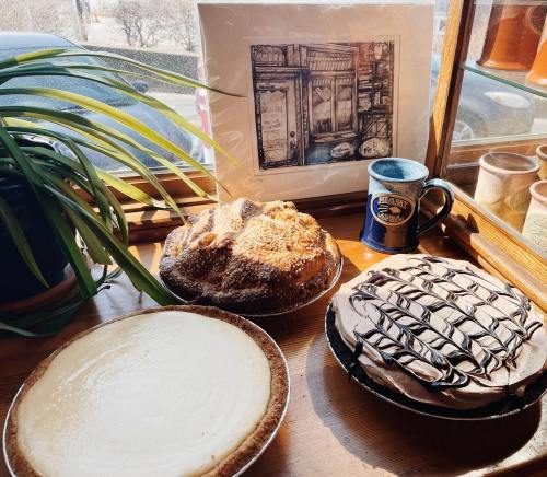 Three delicious desserts on a wooden table: a cheesecake, a crumb-topped cake, and a chocolate pie, with a plant and artwork.