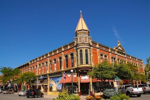 Historic red brick building with a pointed tower, surrounded by trees and shops under a clear blue sky.