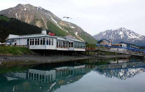 A waterfront restaurant with a wooden deck, surrounded by mountains, reflecting in calm water under a clear sky.
