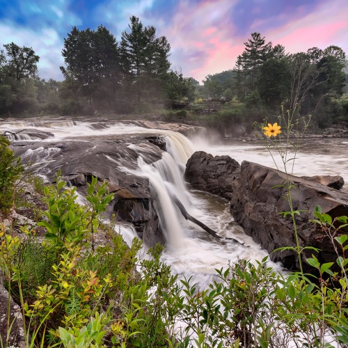 A serene waterfall cascades over rocks, surrounded by lush greenery and a colorful sky at dawn.