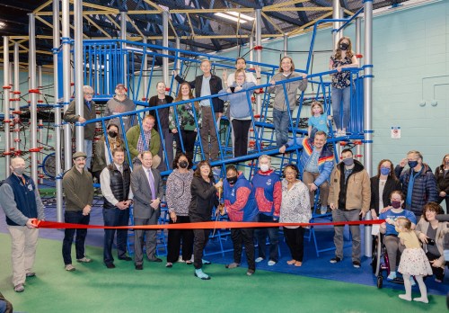 A group of people gathers around a playground structure, celebrating a ribbon-cutting event indoors.