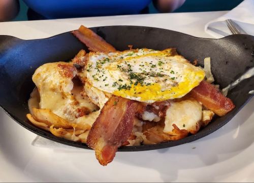 A cast-iron skillet filled with cheesy pasta, crispy bacon, and a fried egg on top, garnished with herbs.