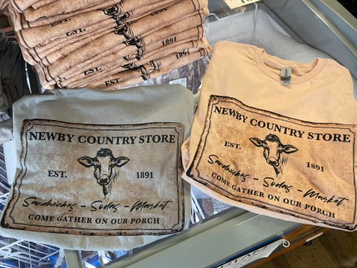 Two t-shirts featuring a vintage-style design with "Newby Country Store" and a cow graphic, stacked on a display.