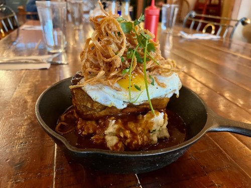 A cast iron skillet holds a dish with a fried egg, crispy onions, mashed potatoes, and savory sauce on a wooden table.