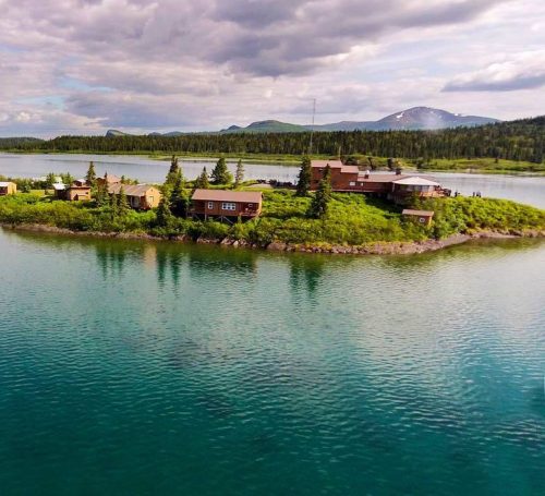 Aerial view of a lush green island with rustic cabins surrounded by calm water and distant mountains under a cloudy sky.