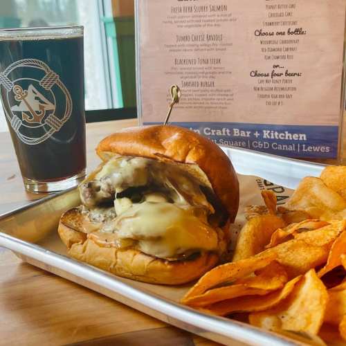 A close-up of a burger with melted cheese and pickles, served with chips and a dark beer in a casual dining setting.