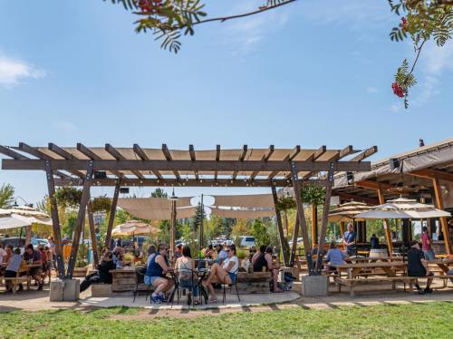 A lively outdoor dining area with people seated at picnic tables under shaded pergolas and umbrellas.