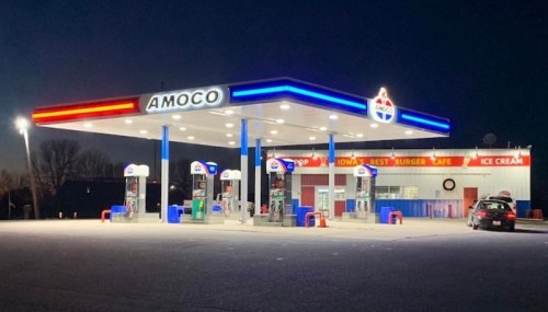 A brightly lit Amoco gas station at night, featuring fuel pumps and a nearby burger cafe with ice cream.