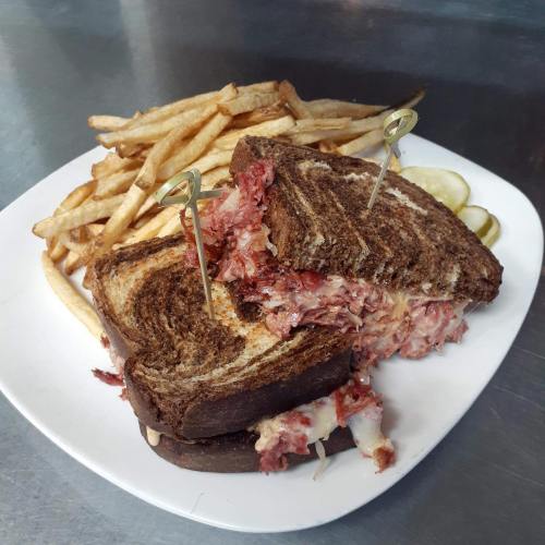 A hearty sandwich with melted cheese and corned beef on rye, served with fries and pickles on a white plate.