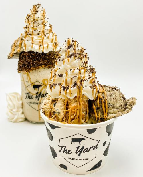 Two decadent milkshakes topped with whipped cream, chocolate drizzle, and cookie pieces in branded cups.