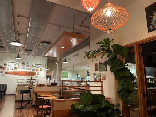 Interior of a restaurant featuring wooden tables, decorative lighting, and green plants, with artwork on the walls.