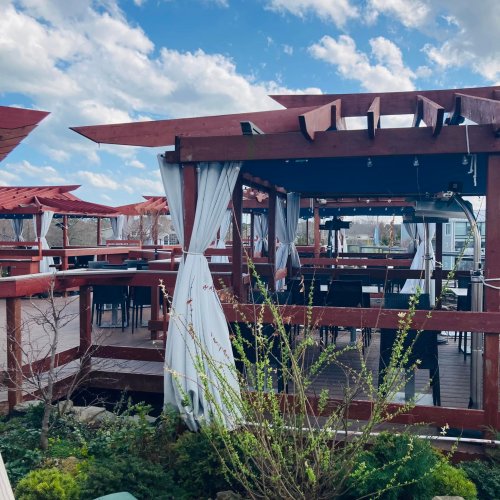 Outdoor dining area with wooden pergolas, draped curtains, and tables under a blue sky with scattered clouds.