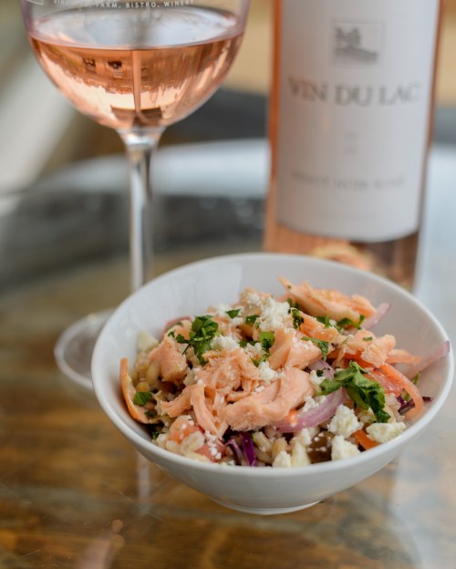 A bowl of salad with salmon, herbs, and cheese, alongside a glass of rosé wine and a bottle in the background.