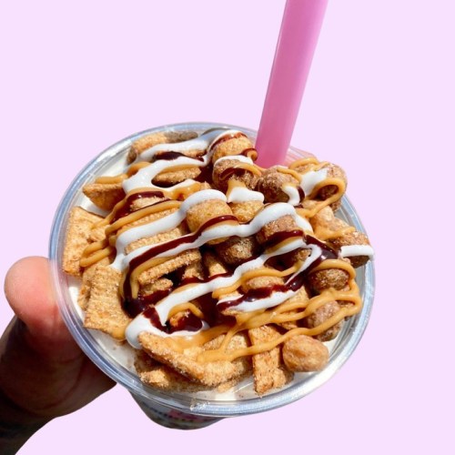 A hand holding a cup filled with ice cream, topped with cereal, chocolate, and peanut butter drizzles.