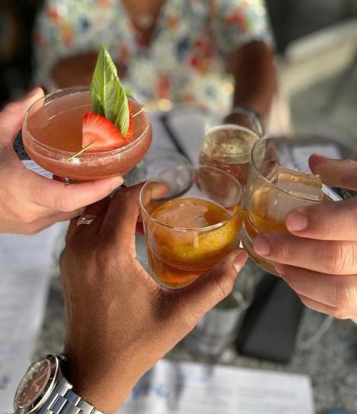 A group of friends toasting with colorful cocktails at a table, featuring garnishes and ice.