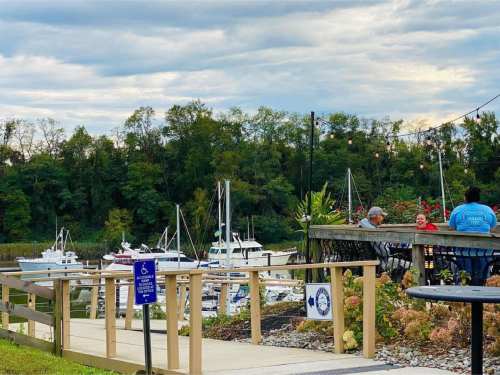 A waterfront scene with boats docked, people sitting at a patio, and trees in the background under a cloudy sky.