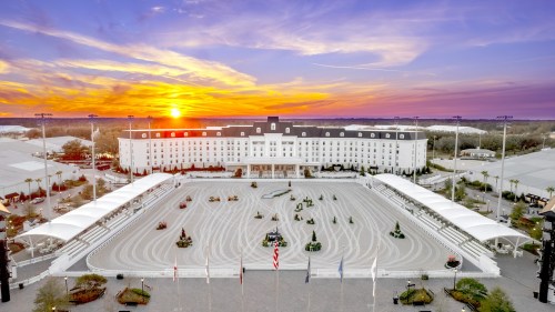 A sunset view of a large equestrian arena with a grand hotel in the background, surrounded by trees and flags.
