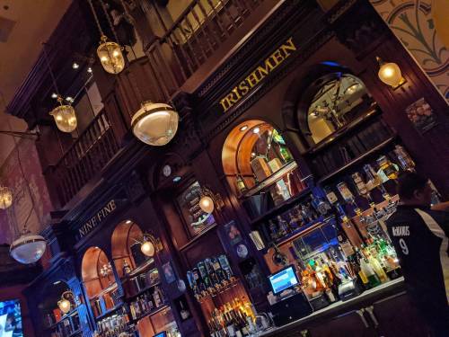 A wooden bar with shelves of liquor, ornate lighting, and a bartender in a sports jersey.