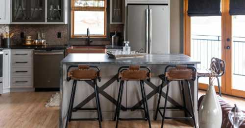 Modern kitchen featuring a large island with three leather bar stools, stainless steel appliances, and wooden accents.