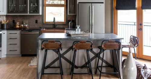 Modern kitchen featuring a large island with three leather bar stools, stainless steel appliances, and wooden accents.